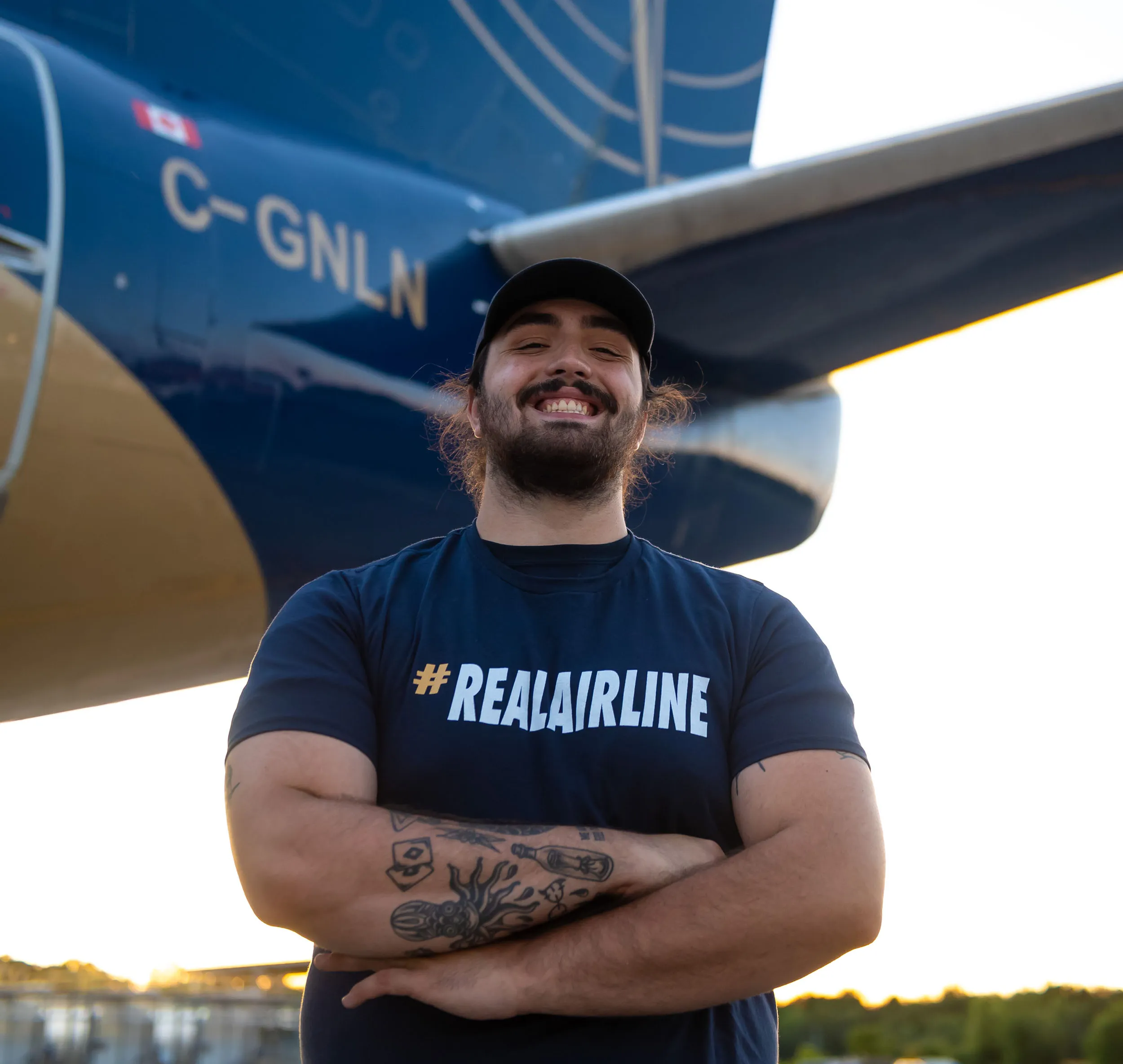 Un homme souriant porte un t-shirt avec le hashtag #REALAIRLINE, debout devant un avion aux couleurs bleu et or.