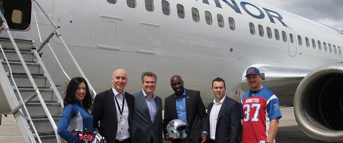 Group of six people, including cheerleaders and team members, pose with a football helmet near a plane marked "MINOR."