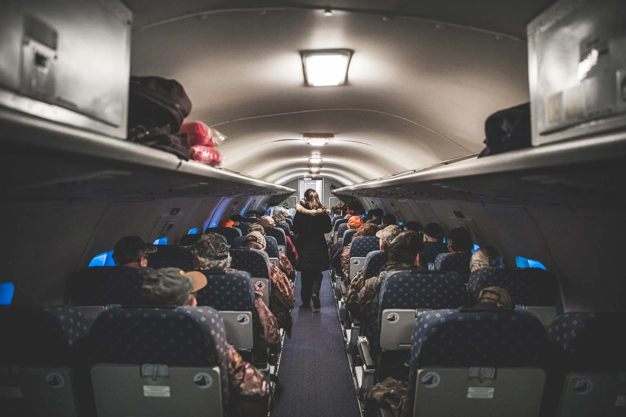 Intérieur d'un avion avec des passagers en vêtements de camouflage assis sur des sièges. Une hôtesse de l'air se dirige vers l'arrière.