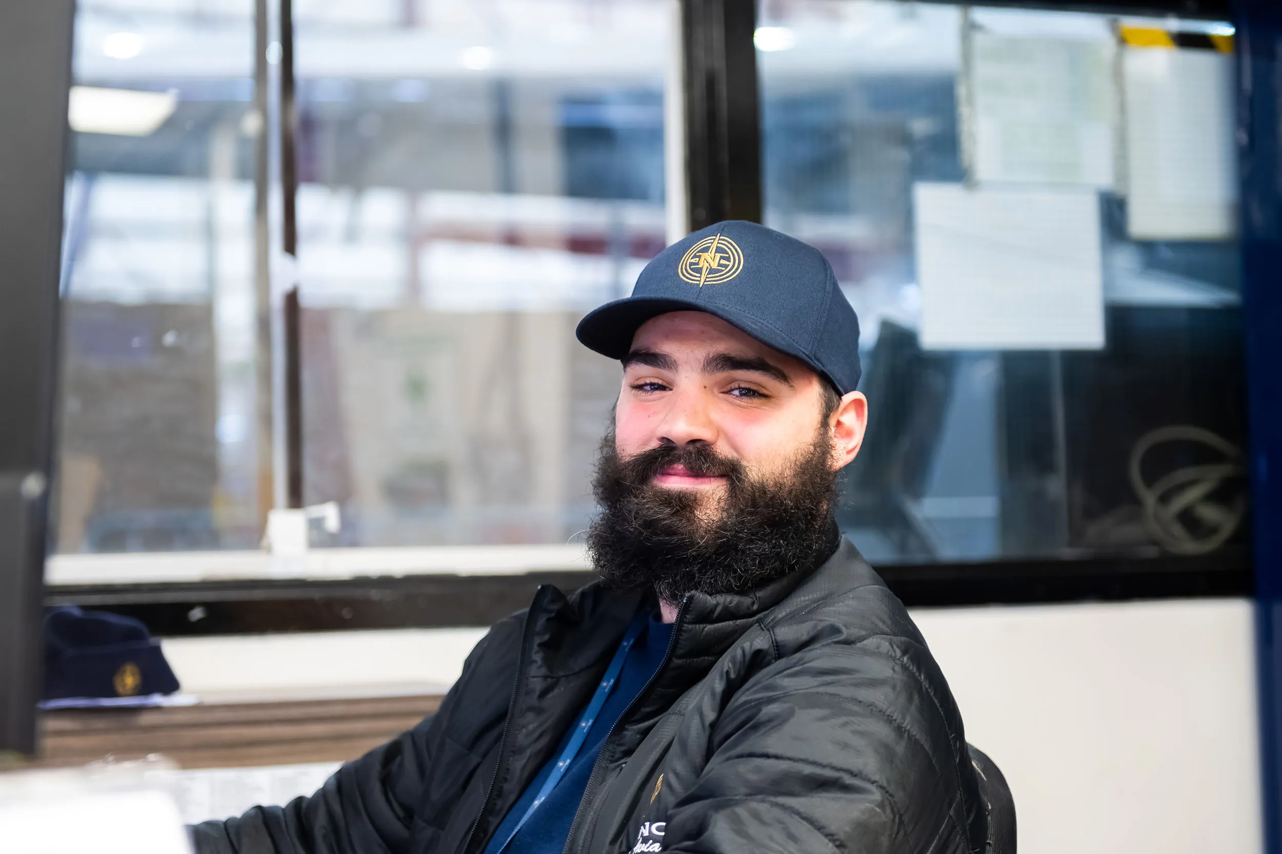 Un homme avec une barbe et une casquette sourit depuis son poste de travail, entouré de documents et d'un environnement de bureau.