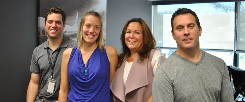 Quatre personnes souriantes se tiennent côte à côte dans un bureau moderne, avec des fenêtres laissant passer la lumière naturelle.