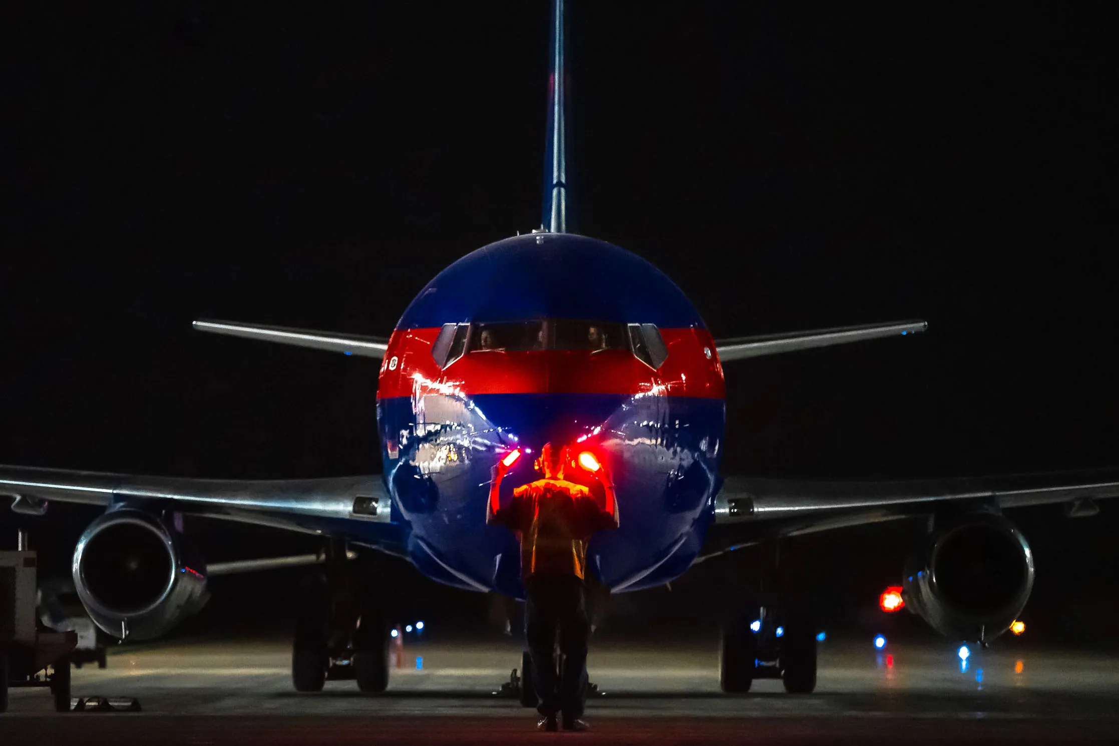 Un avion de ligne aux couleurs vives est stationné sur le tarmac, un technicien avec une lampe frontale s'approche pour effectuer des vérifications.