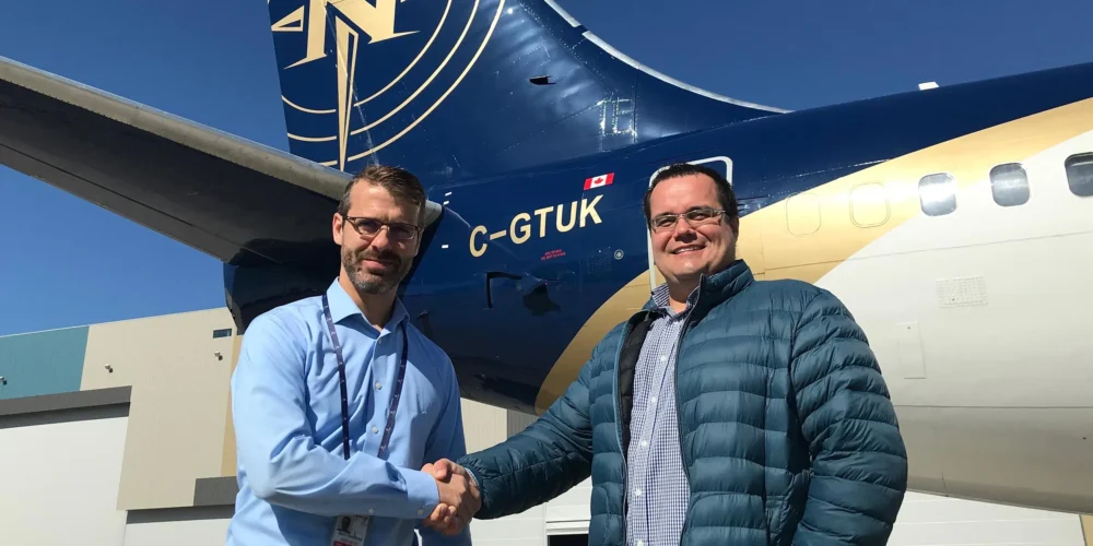 Deux hommes se serrent la main devant un avion, l'un porte une chemise bleue et l'autre une veste matelassée. L'avion est marqué C-GTUK.