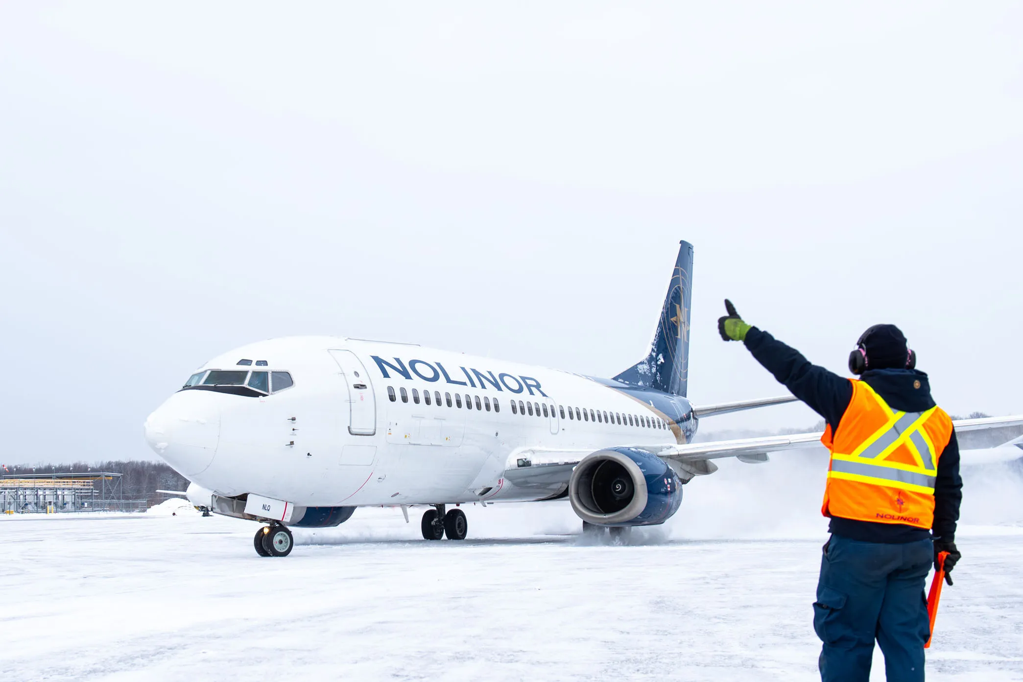 Un avion de la compagnie Nolinor se prépare à décoller sur une piste enneigée, tandis qu'un agent au sol dirige le trafic avec un geste de la main.