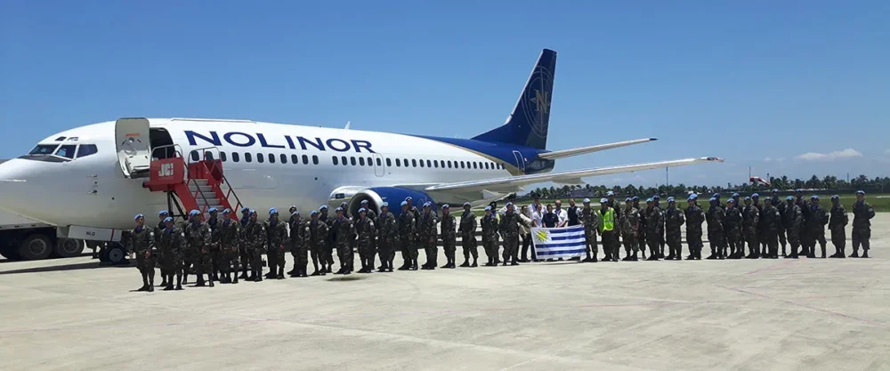 Des soldats en uniforme se tiennent en rang devant un avion Nolinor, avec un drapeau en arrière-plan. Ciel dégagé et ambiance festive.