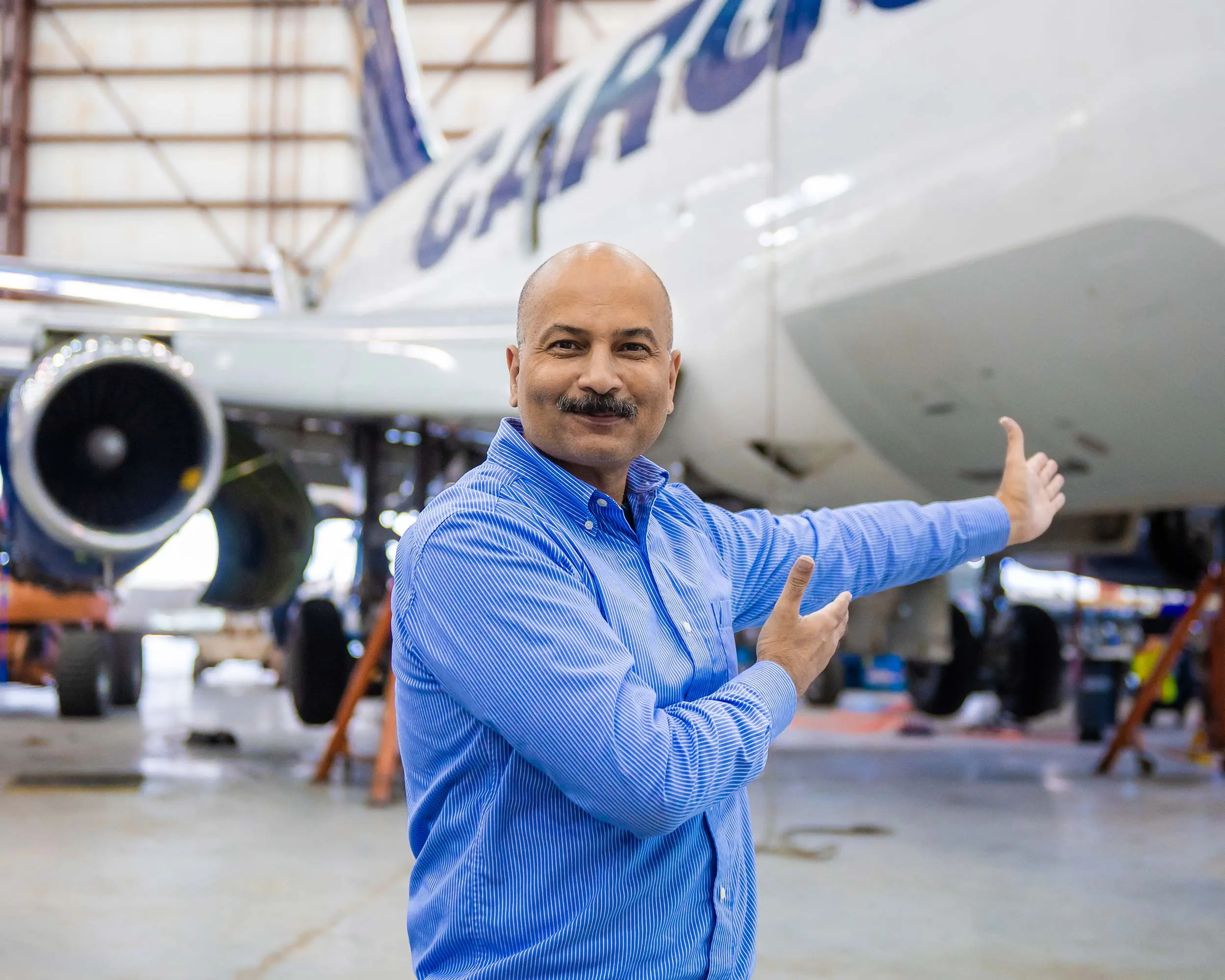 Un homme souriant montre un avion de transport en arrière-plan, dans un hangar d'entretien aéronautique. Il porte une chemise bleue.