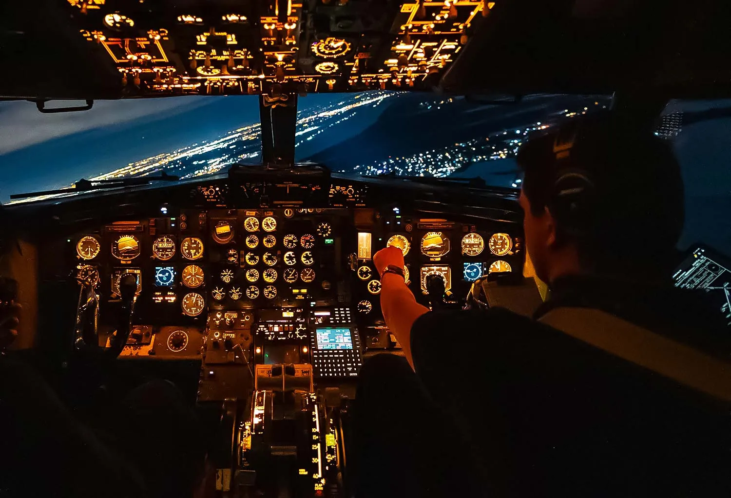 A pilot operates an aircraft cockpit at night, illuminated by instrument lights, with city lights visible outside.