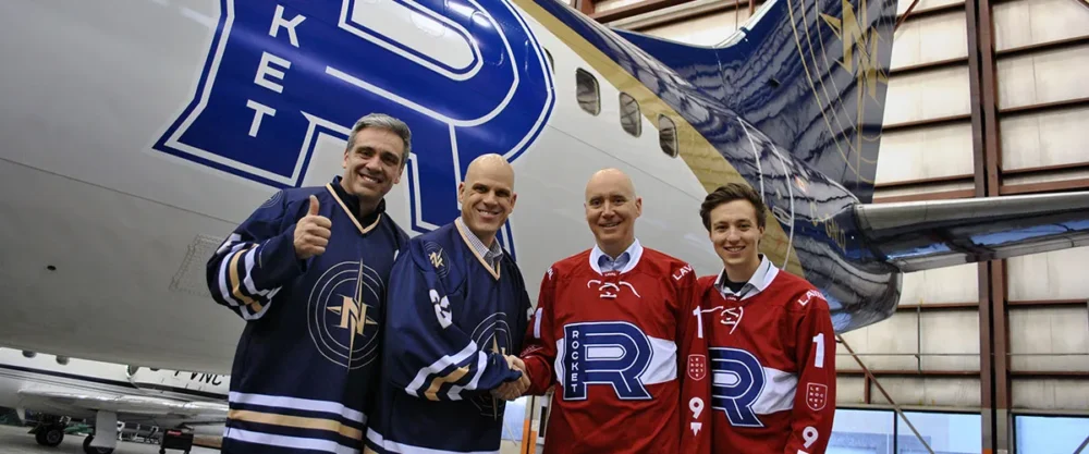 Quatre hommes posent devant un avion, souriant et portant des maillots de hockey. Deux maillots sont rouges avec le logo "Rocket".