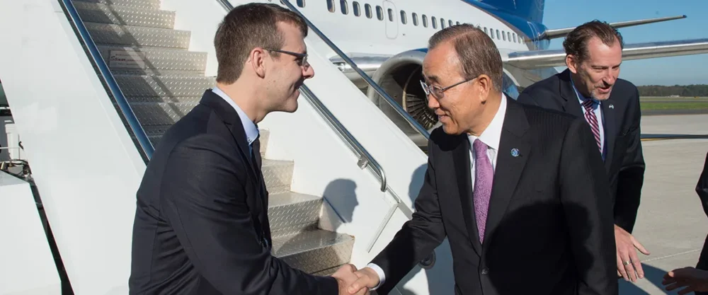Deux hommes se serrent la main sur un tarmac d'aéroport, un homme en costume souriant et un autre avec des lunettes, accompagné d'un troisième homme en arrière-plan.
