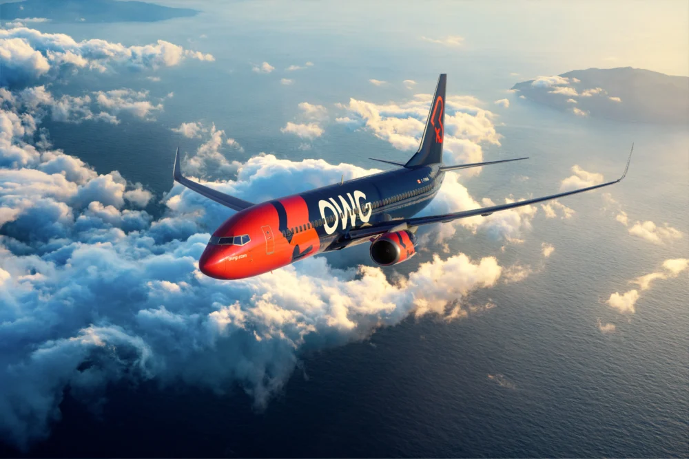 Un avion de ligne orange et bleu survole des nuages au-dessus de l'océan, avec des montagnes en arrière-plan.