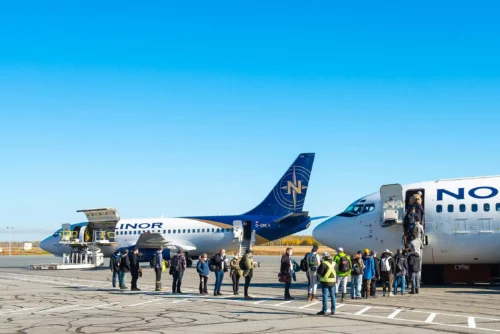 Des passagers descendent d'un avion sur le tarmac, tandis qu'un autre avion est stationné à proximité sous un ciel bleu clair.