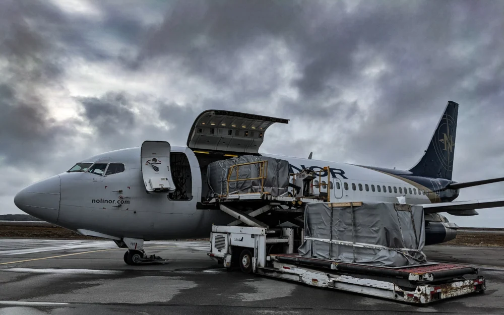 Un avion de transport de marchandises est garé sur le tarmac, avec une porte ouverte et des palettes de fret en cours de chargement. Le ciel est nuageux.