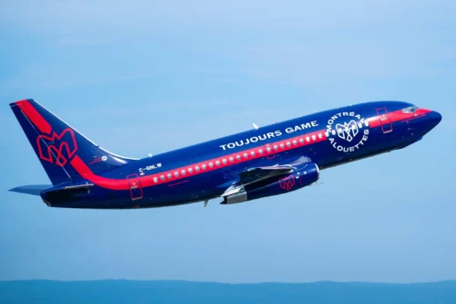Un avion de ligne bleu et rouge décolle, affichant le logo de Montréal et le slogan "TOUJOURS GAME" sur le fuselage.
