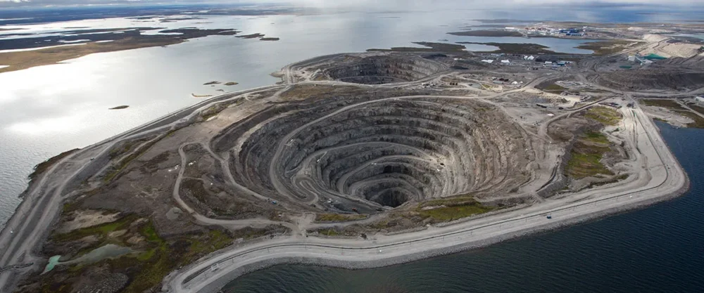 Un vaste site minier avec une grande fosse circulaire entourée d'eau, des routes et des infrastructures visibles au loin.