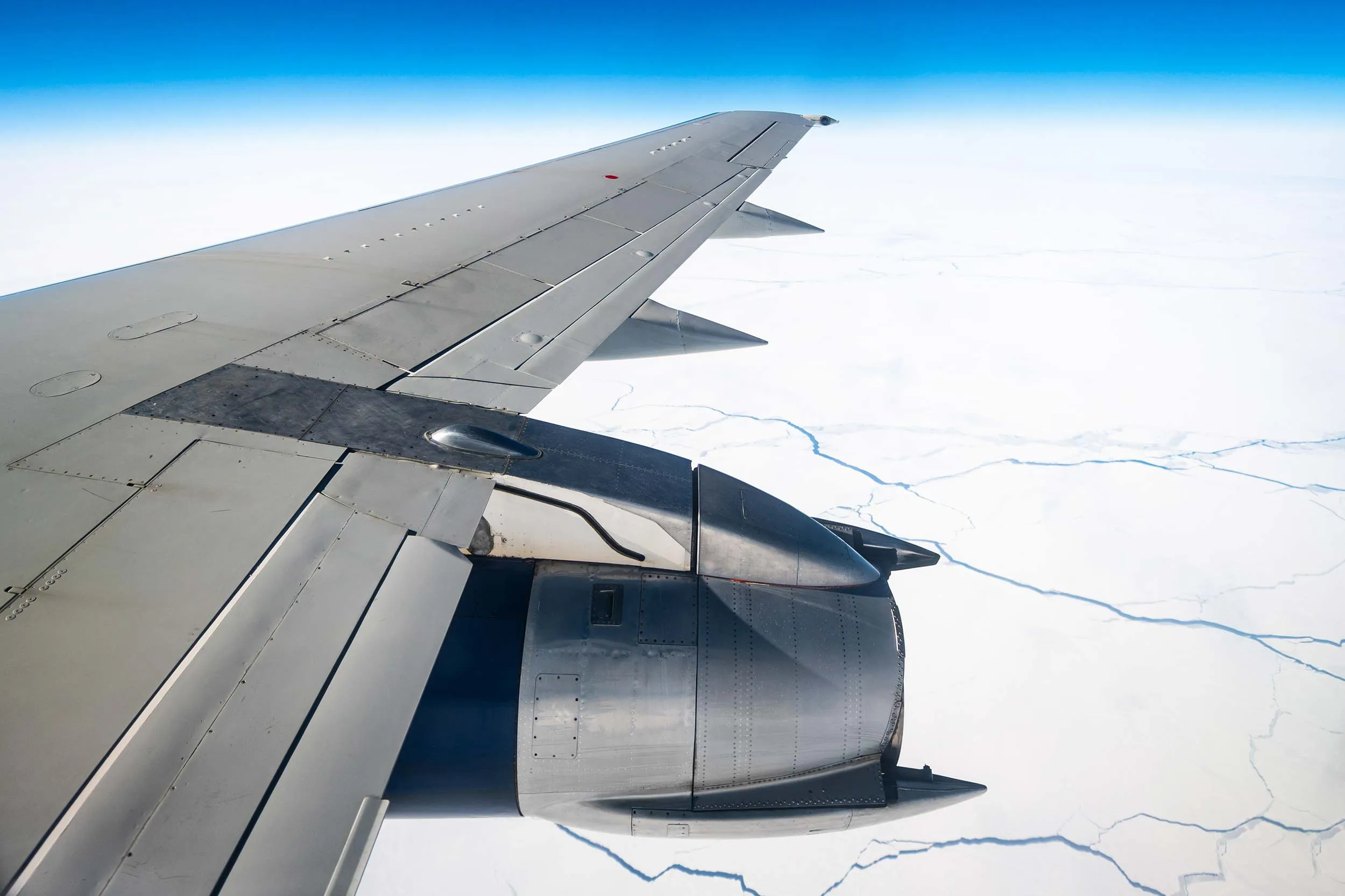 Vue de l'aile d'un avion survolant un paysage enneigé, avec des fissures dans la glace visibles en dessous. Ciel dégagé et bleu.