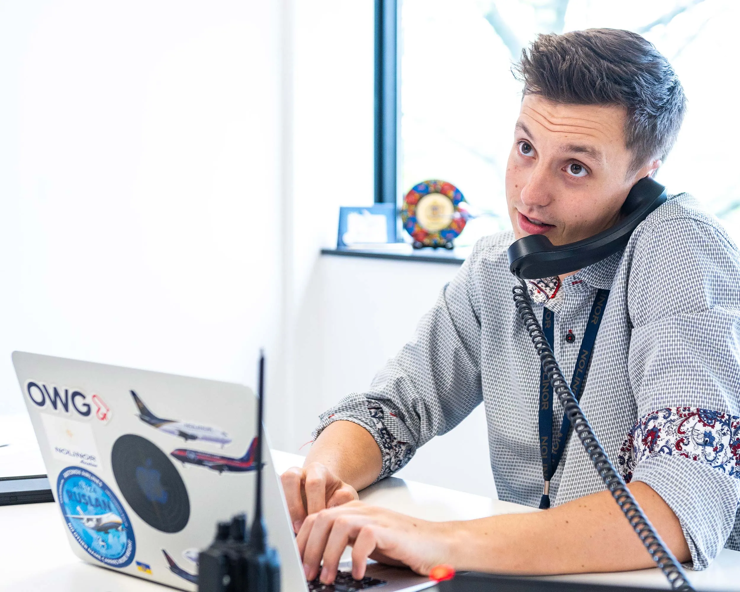 Un jeune homme au bureau parle au téléphone tout en tapant sur son ordinateur portable décoré d'autocollants. Une fenêtre en arrière-plan laisse entrer la lumière.