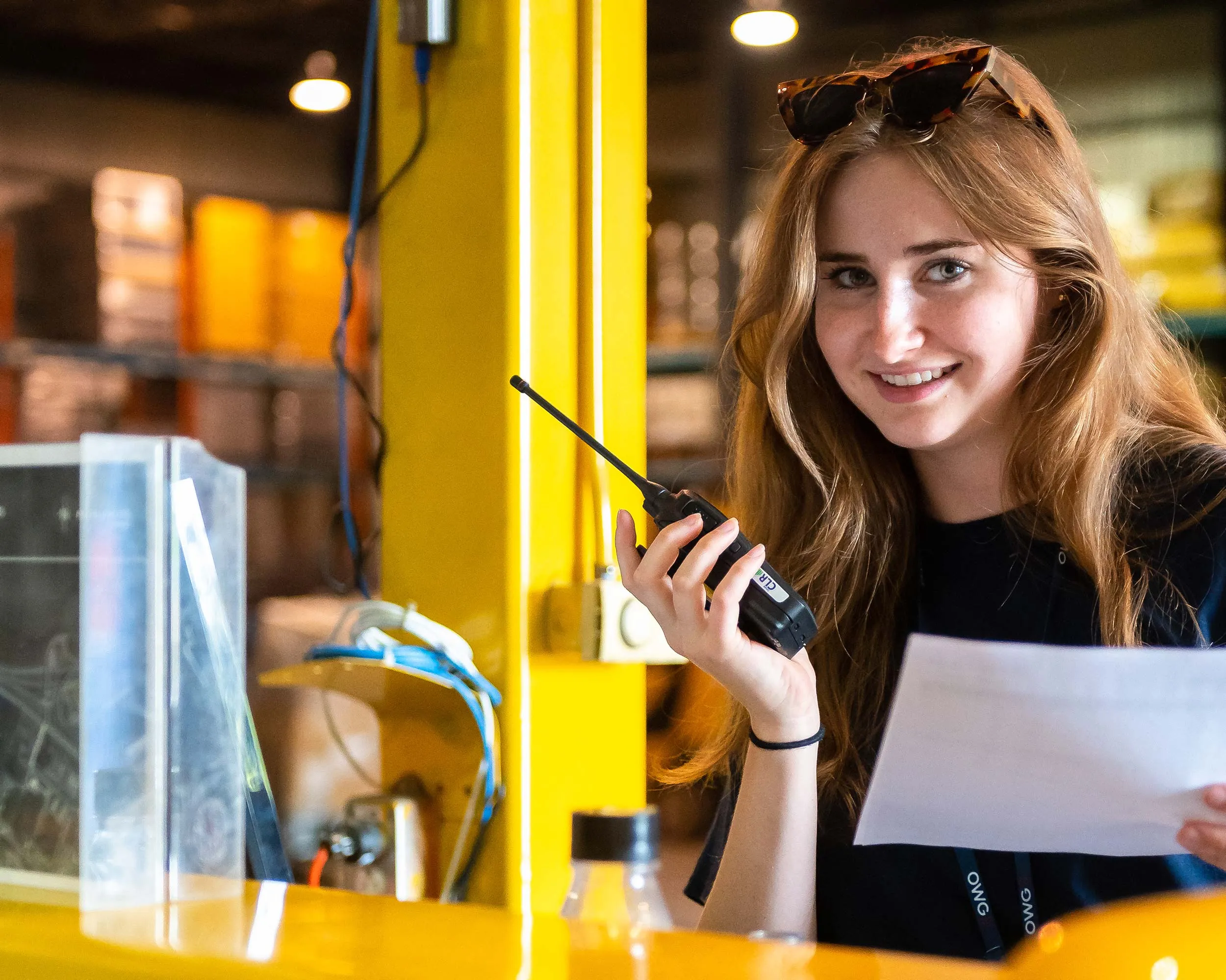 Une jeune femme souriante tient une radio tout en consultant un document. Elle se trouve dans un environnement de travail lumineux avec des étagères en arrière-plan.