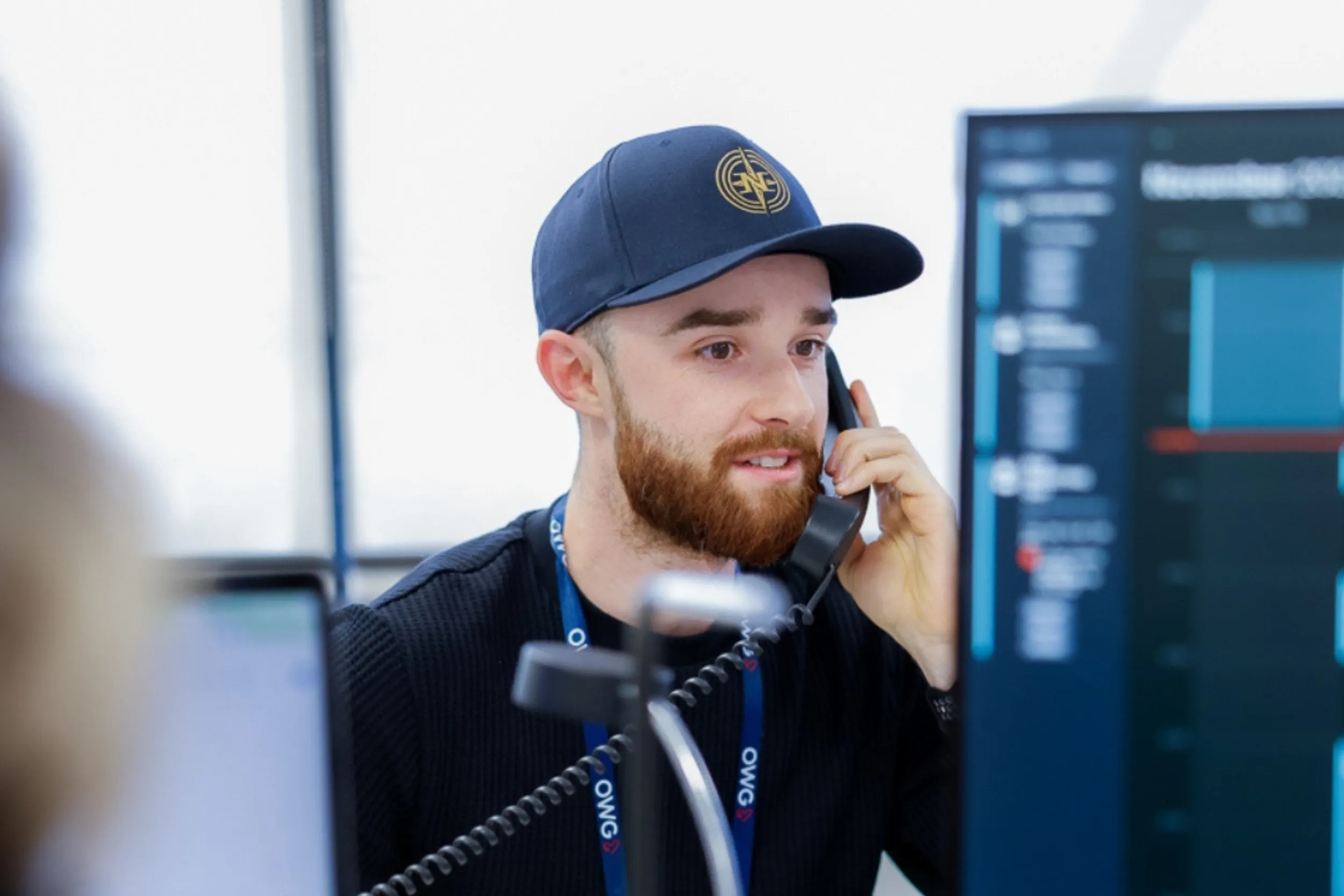 Un homme avec une casquette parle au téléphone, assis devant un écran d'ordinateur affichant des données. Il semble concentré et engagé dans une conversation.