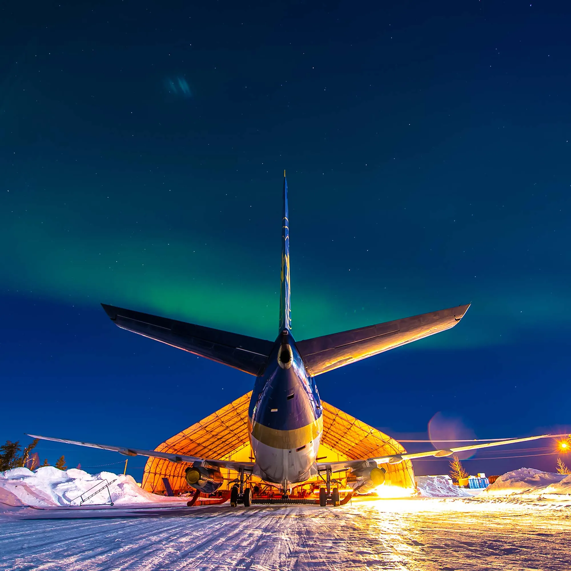 Un avion est stationné devant un hangar illuminé sous les aurores boréales, créant une atmosphère magique dans un paysage enneigé.
